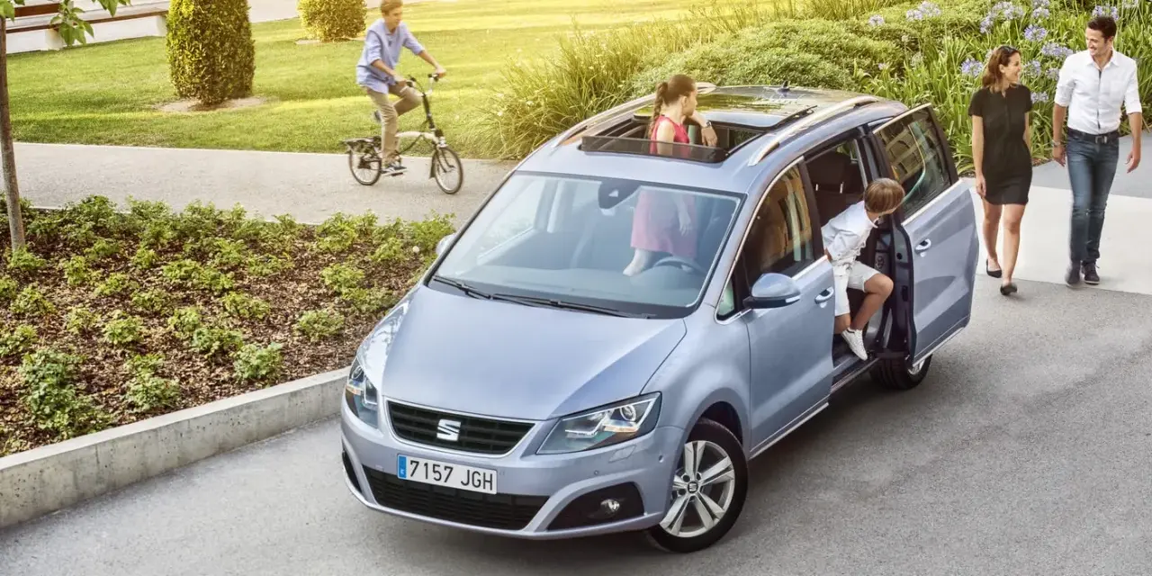 Familienauto mit zwei Kindern, die ein- und aussteigen. Ein Junge fährt Fahrrad im Hintergrund.