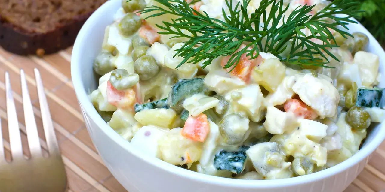 Ein Schüsselchen Olivier Salat, garniert mit Dill, neben einer Scheibe dunklem Brot und einer Gabel.