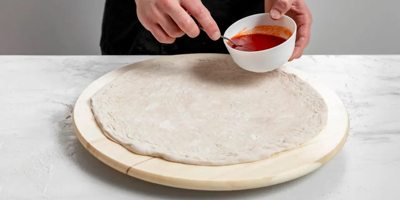 Przygotowanie ciasta na pizzę bez drożdży. Dłonie nabierają sos pomidorowy z miseczki, by rozprowadzić go na okrągłym spodzie.