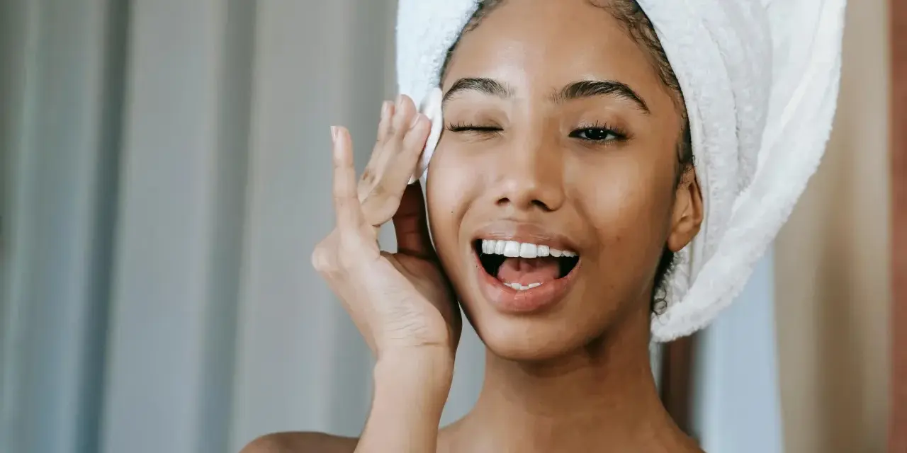 Une jeune femme avec une serviette sur la tête, appliquant un soin sur son visage. Elle semble heureuse, peut-être grâce aux bienfaits du collagène ou de l'acide hyaluronique.
