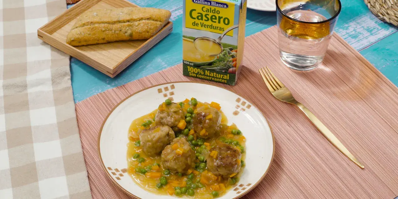 Albondigas a la jardinera en un plato blanco, acompañadas de un pan rústico, un vaso de agua y un caldo de verduras.