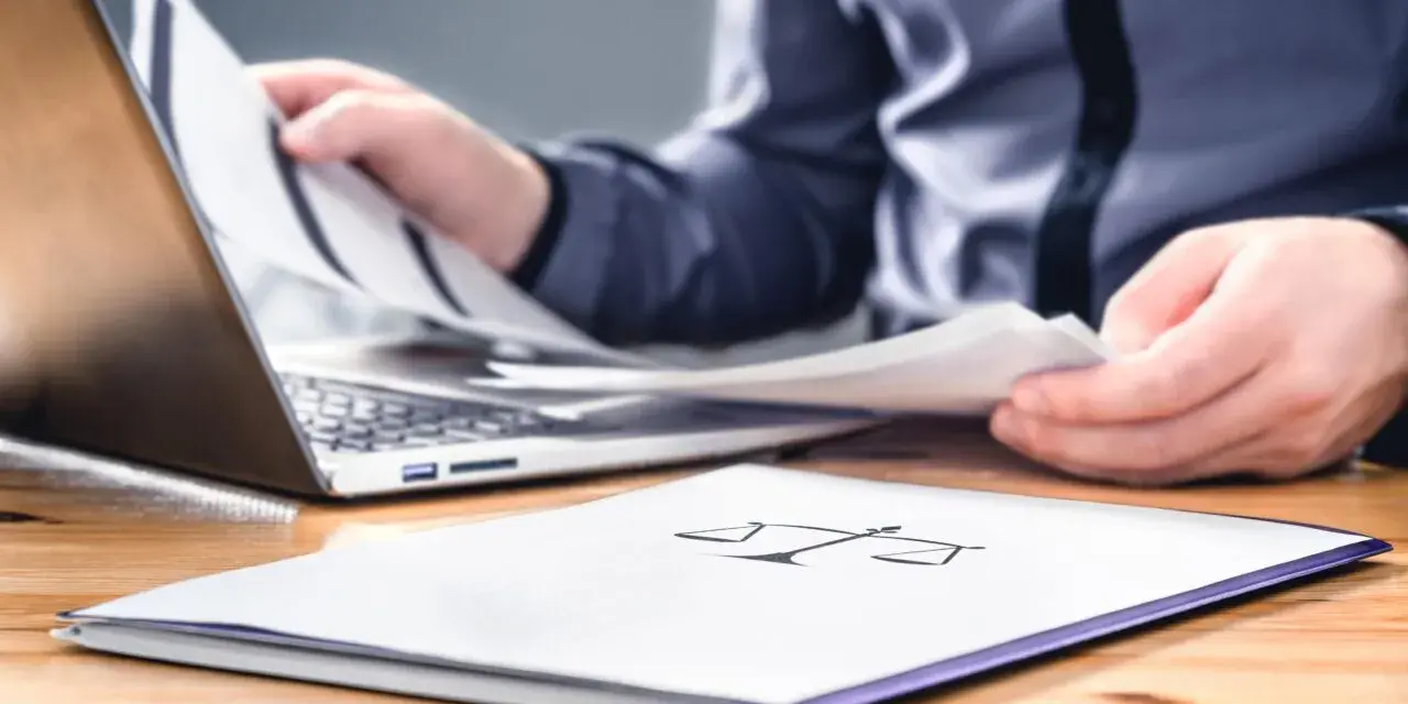 Osoba przegląda dokumenty przy laptopie. Na pierwszym planie folder z symbolem wagi, sugerujący sprawy związane z prawem i potencjalną masą upadłościową.