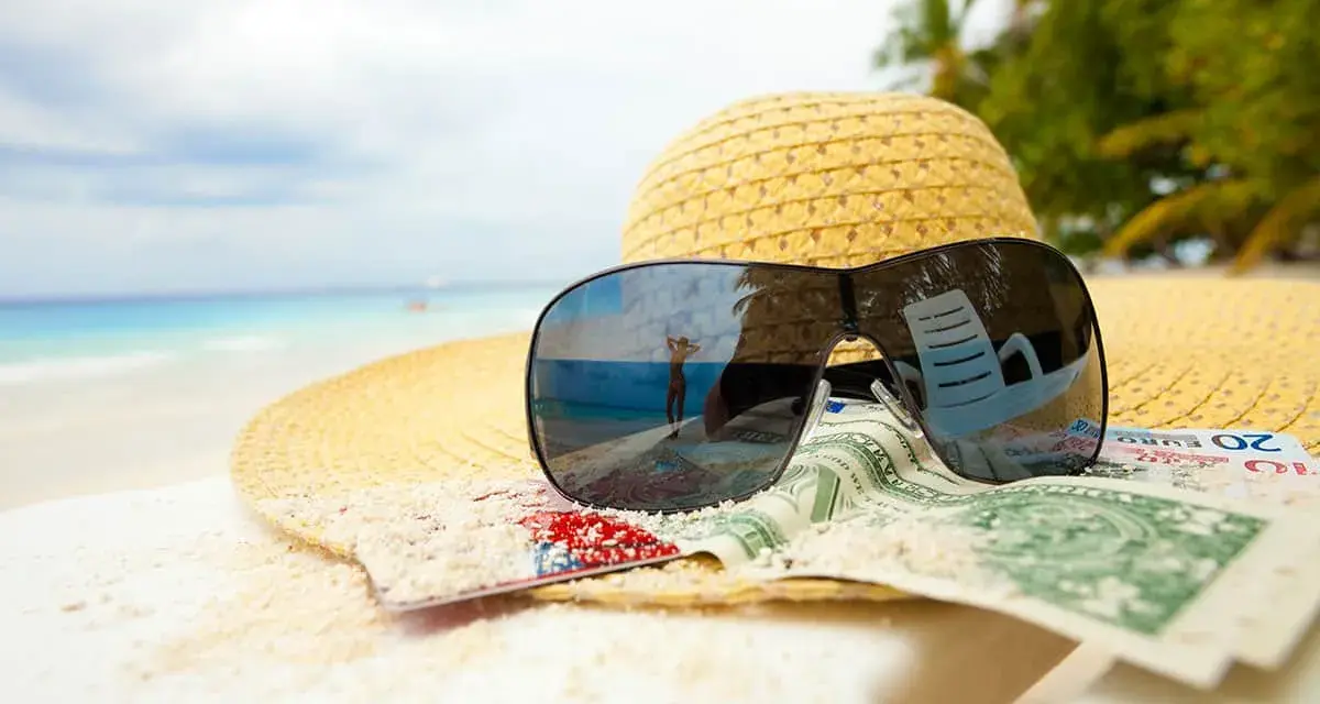 Sonnenbrille, Hut und Geld am Strand. Der Spiegel der Brille zeigt eine Person, die den Urlaub genießt. Ein Symbol für den **Urlaubsgeldanspruch**.