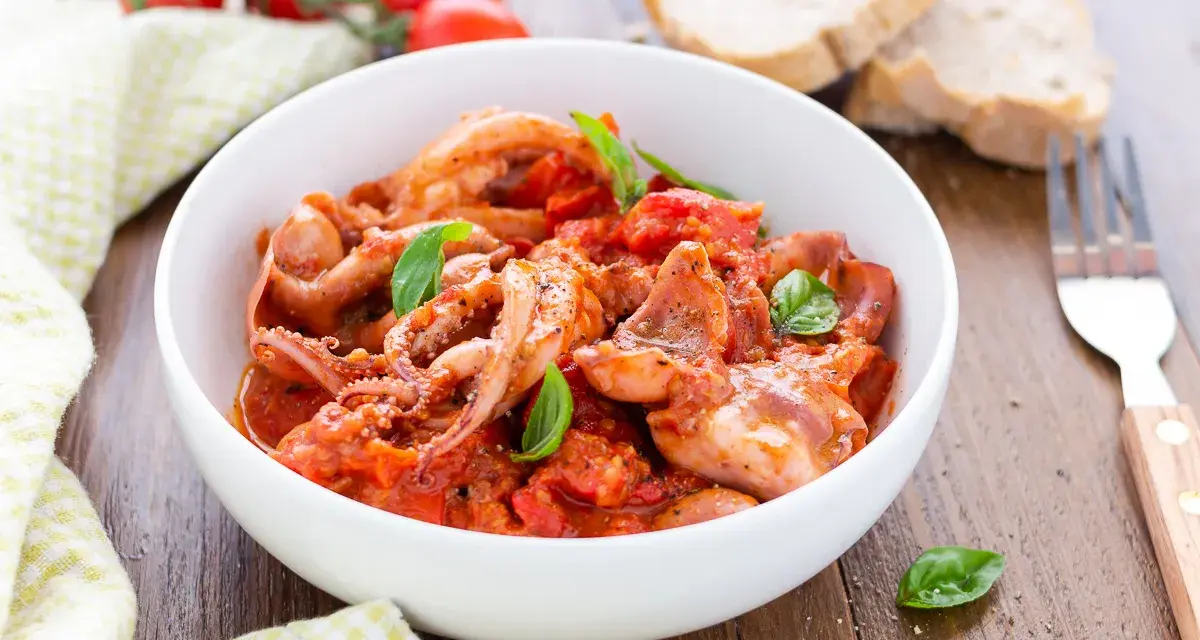 Totani in umido con pomodoro e basilico, serviti in una ciotola bianca, con pane e forchetta sullo sfondo.