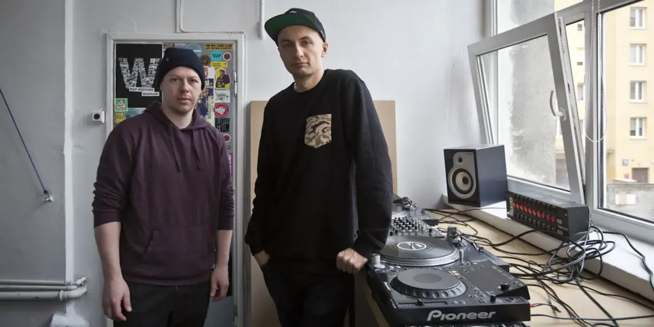 Dwóch mężczyzn przy sprzęcie DJ-skim. Jeden w bordowej bluzie z kapturem, drugi w czarnej bluzie z naszywką. To ich sposób na pokazanie, co to stereo.