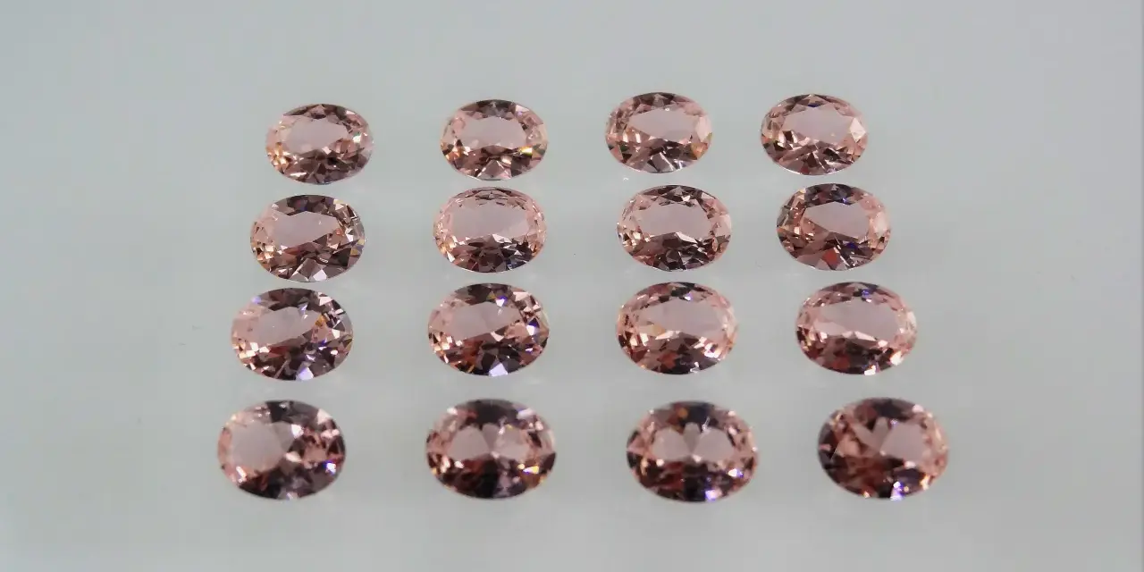 Sedici pietre di morganite ovali, rosa pesca, disposte in file ordinate. Le loro sfaccettature riflettono la luce, mostrando le belle morganite proprietà.