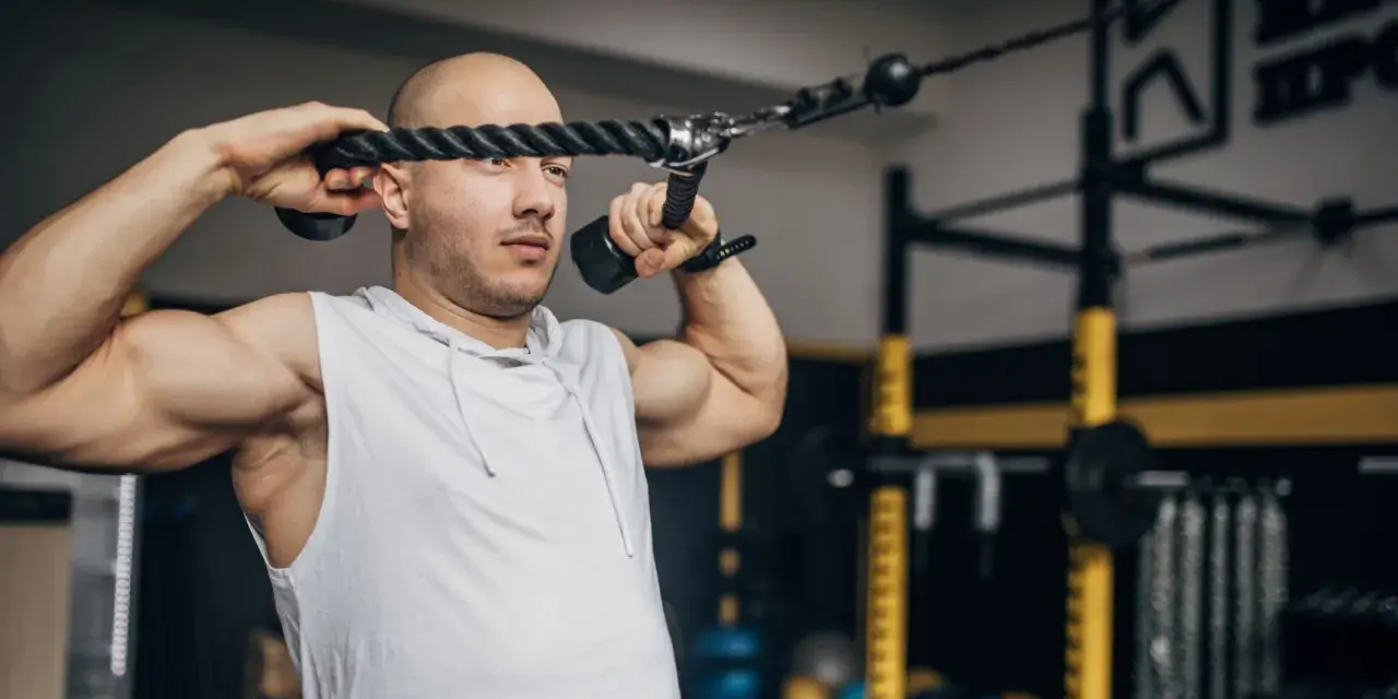 Man med rakad skalle utför face pulls med ett rep i ett gym.