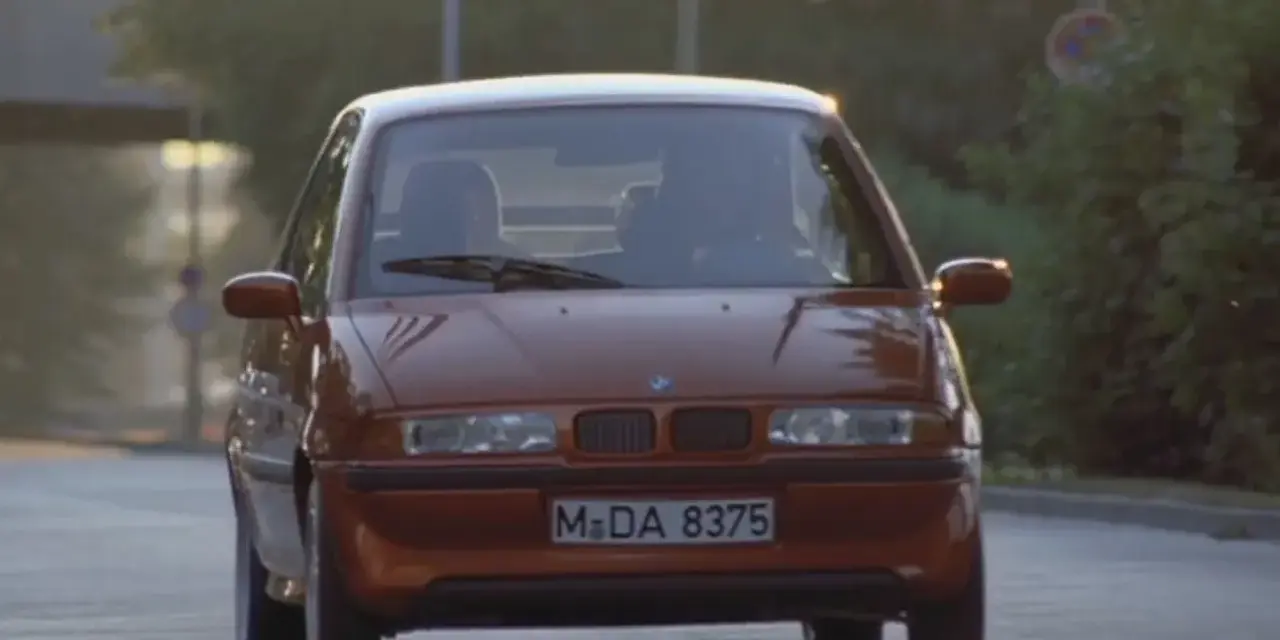 Pomarańczowy BMW E1 jedzie po drodze, światła odbijają się od karoserii.