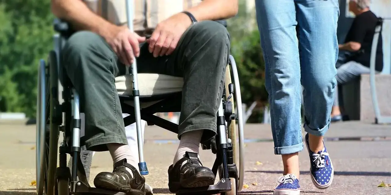Osoba na wózku inwalidzkim z laską, obok niej idzie osoba w jeansach.