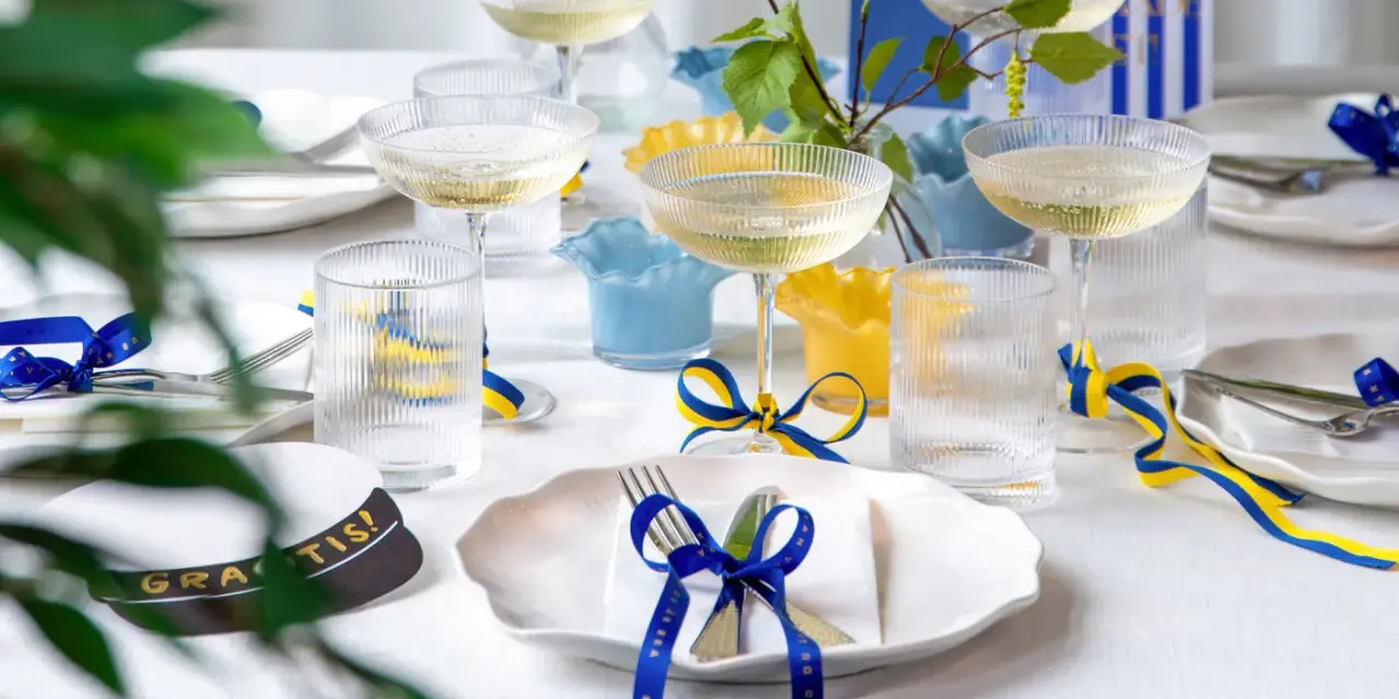 Festligt dukat bord med champagneglas, blå och gula band som knyts i en blå rosett, samt en studentmössa.