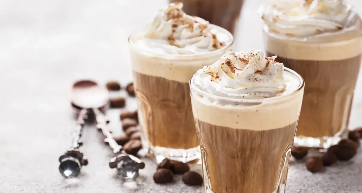 Tre bicchieri di deliziosa crema al caffè, guarniti con panna montata e cacao. Chicchi di caffè e cucchiaini decorati completano la scena.
