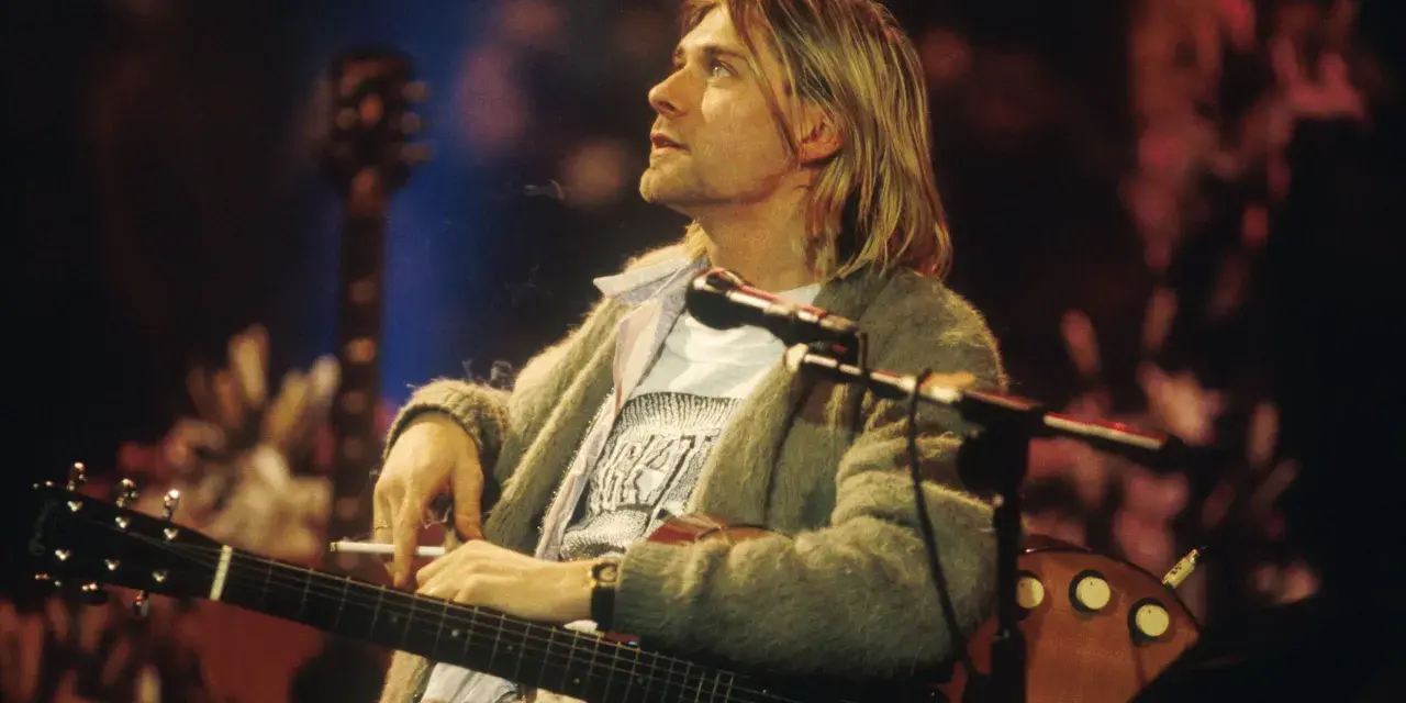 Kurt Cobain z papierosem w dłoni, grający na gitarze. Jego muzyka to część 100 najlepszych piosenek wszechczasów.
