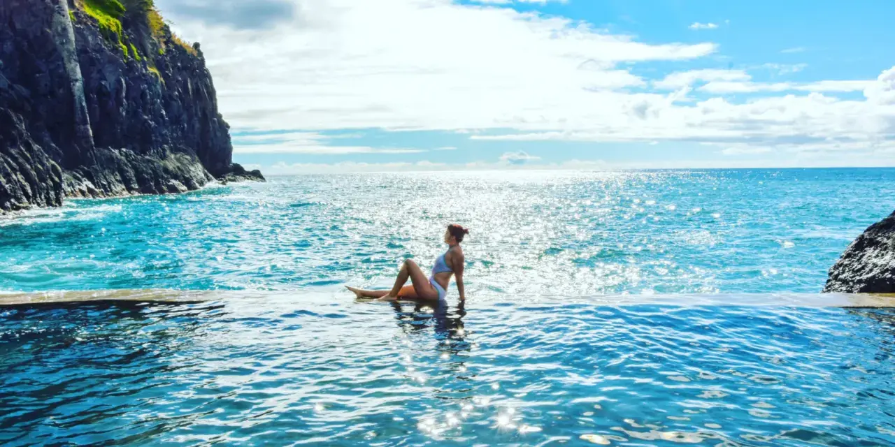 Madera, listopad. Słoneczna pogoda zachęca do kąpieli w basenie z widokiem na ocean. Kobieta w białym bikini relaksuje się na brzegu.