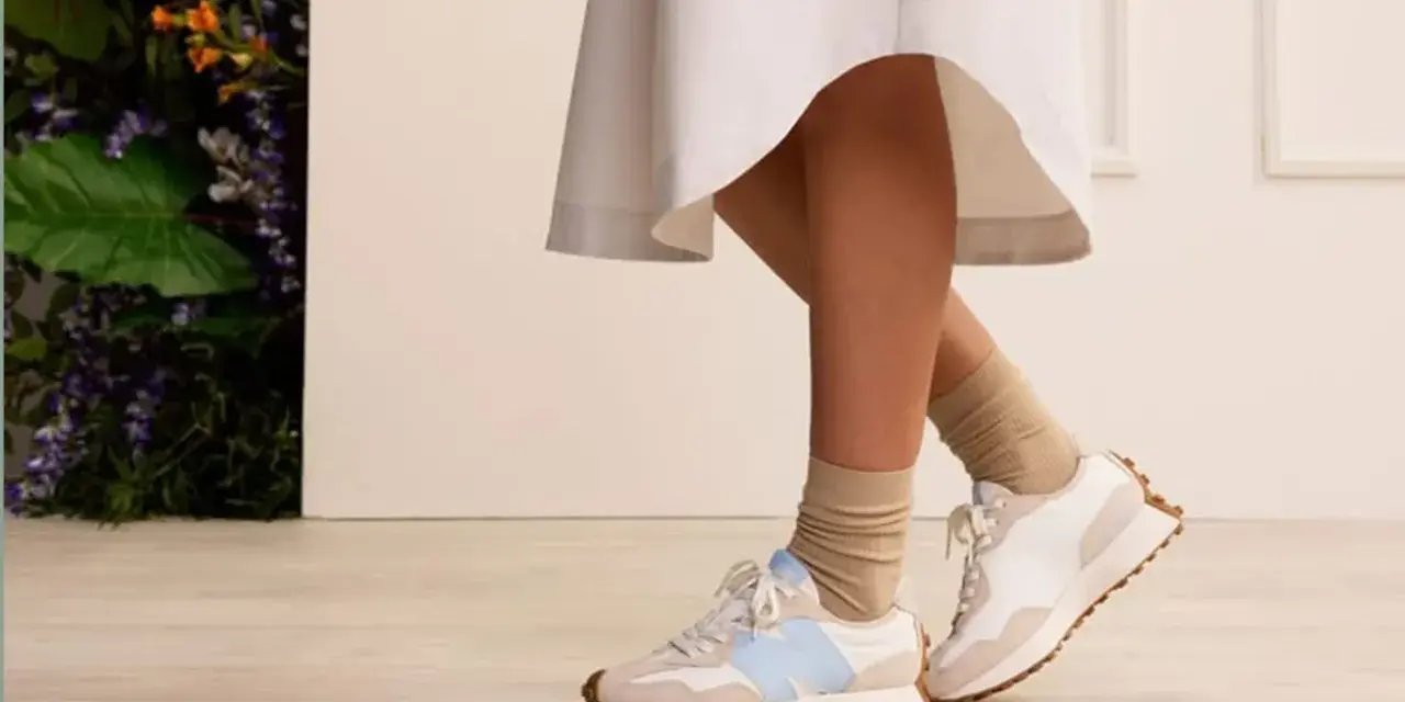 Elegancka sukienka i sportowe buty tworzą stylowy kontrast. Nogi w beżowych skarpetkach i białych sneakersach z błękitnym logo.