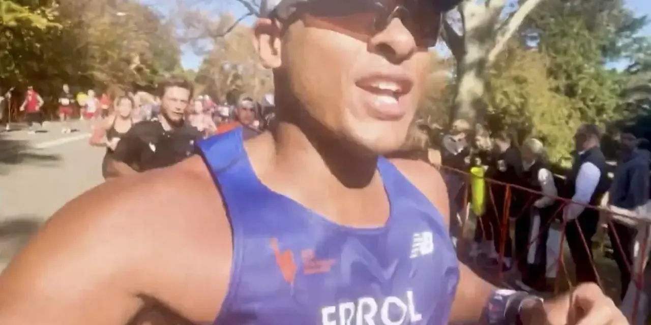 En löpare i blått linne, med texten "ERROL", kämpar på för att springa marathon. Publiken hejar längs banan.