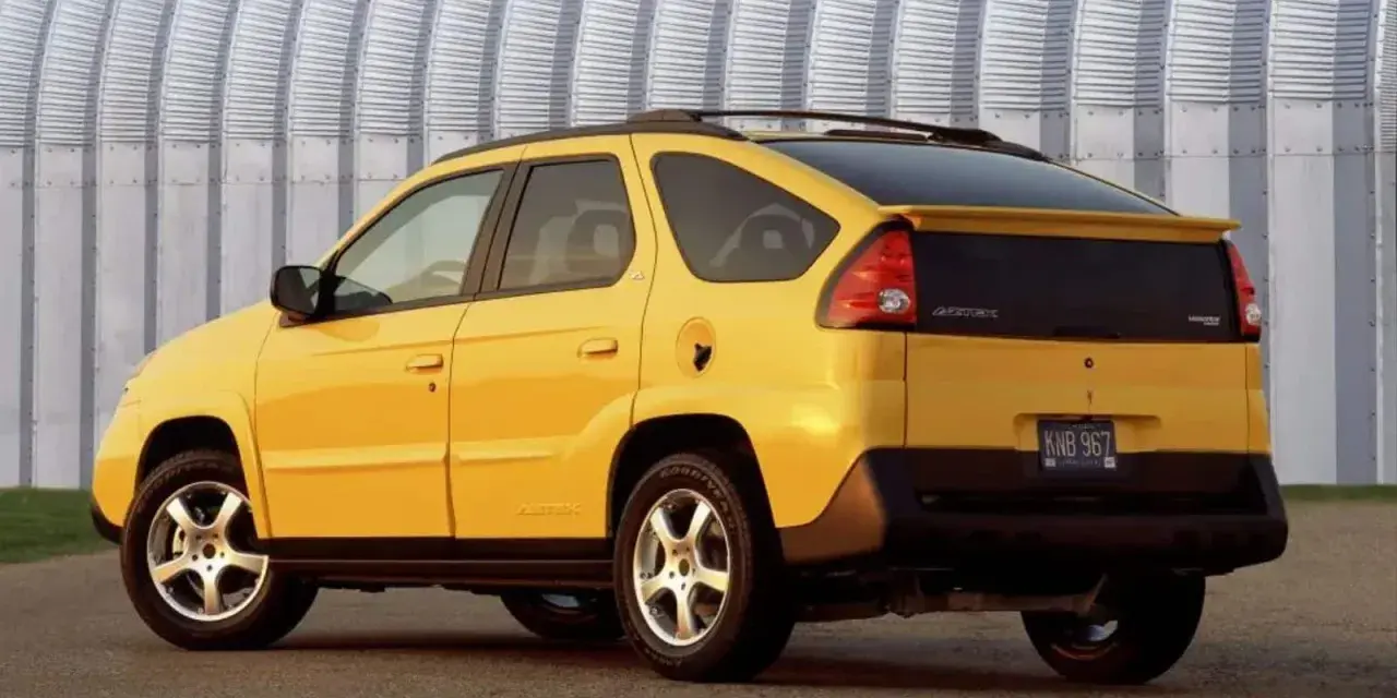 Żółty Pontiac Aztek z charakterystycznym tyłem i spojlerem, zaparkowany przed falistą, metalową ścianą.