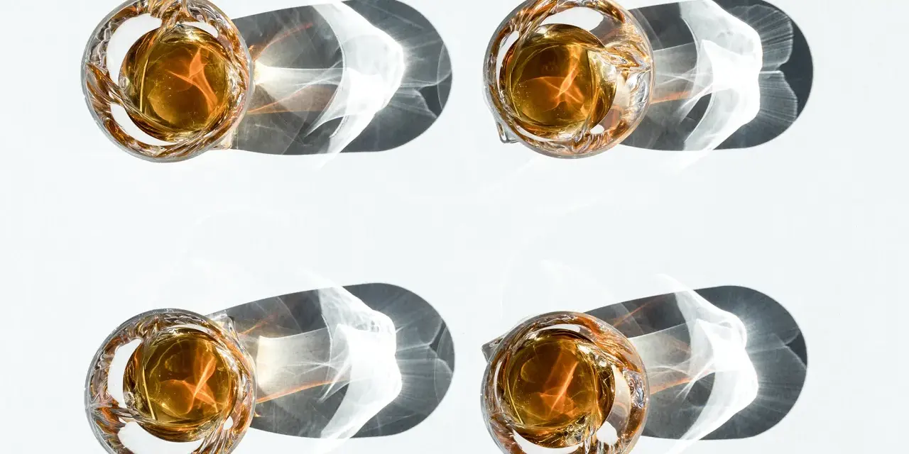 Vier Gläser mit bernsteinfarbenem Whisky werfen dramatische Schatten. Ein visueller Vergleich, der den Unterschied Scotch und Bourbon verdeutlicht.