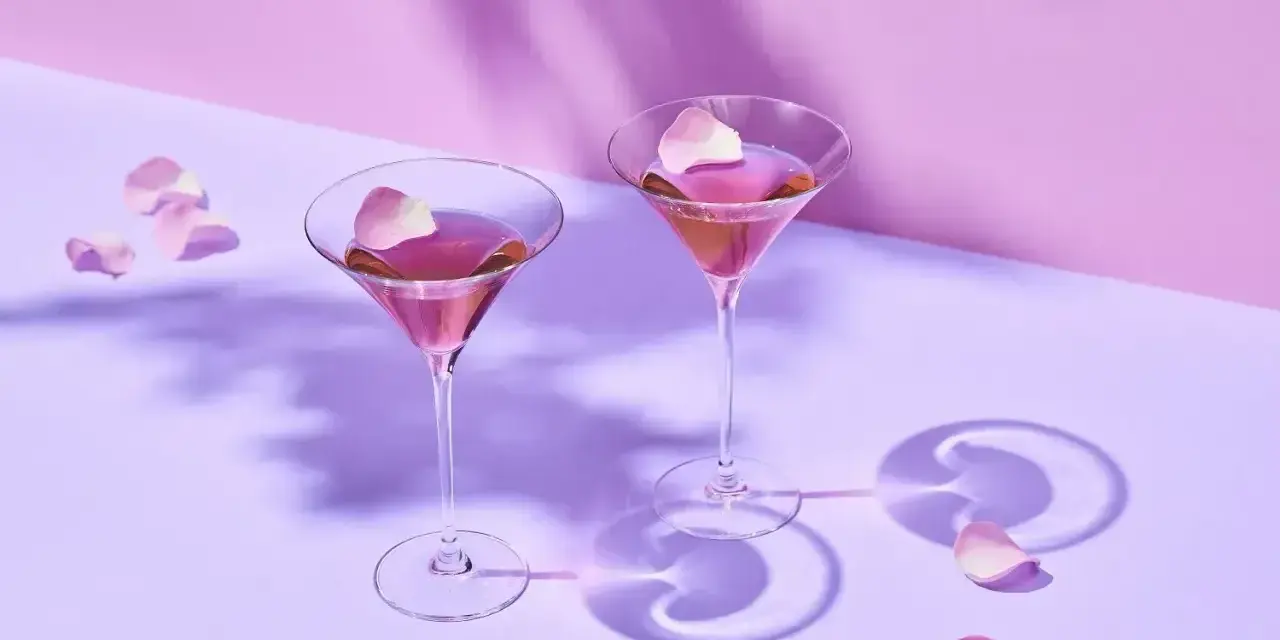 Elegancki Martini przepis: dwa kieliszki z różowym drinkiem, ozdobione płatkami róż, na fioletowym tle.