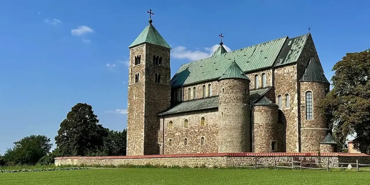 Kamienny zamek w Tumie z zielonym dachem, otoczony murem i zieloną łąką, pod błękitnym niebem.