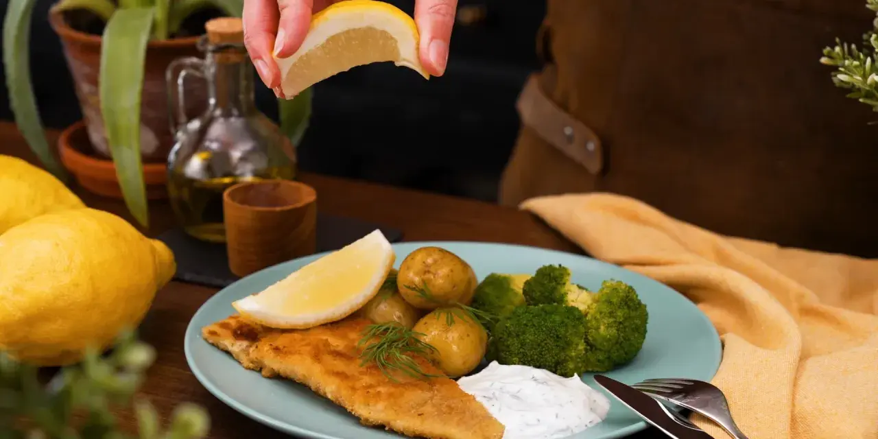 En hand pressar citron över en tallrik med panerad torsk, potatis, broccoli och en klick sås.