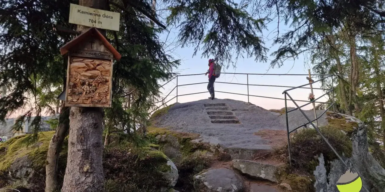 Widok na szczyt Kamienna Góra (704 m). Turystka podziwia panoramę, a obok drzewa ozdobiona kapliczka.