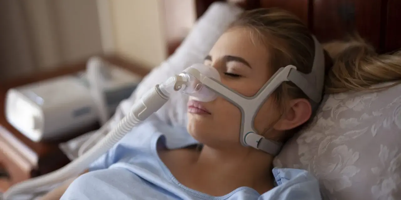 Kobieta śpi w masce CPAP, która pomaga w leczeniu bezdechu sennego. Urządzenie zapewnia ciągłe doprowadzanie powietrza, łagodząc objawy.