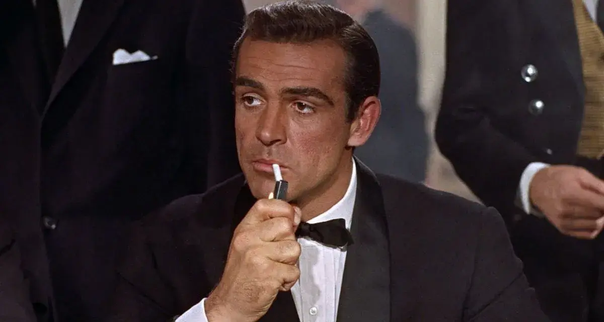 James Bond, elegante in smoking, accende una sigaretta con un'aria pensierosa.