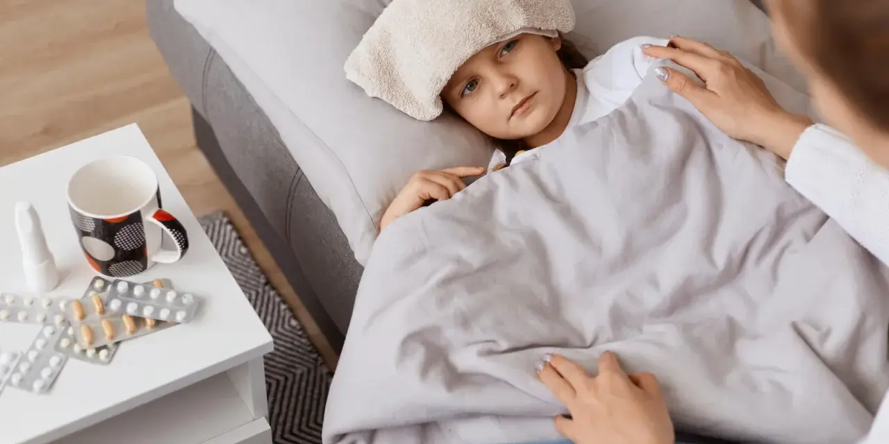 Chora dziewczynka z okładem na głowie leży pod kocem. Obok leki i kubek. Mama czuwa, zapewniając opiekę na dziecko.
