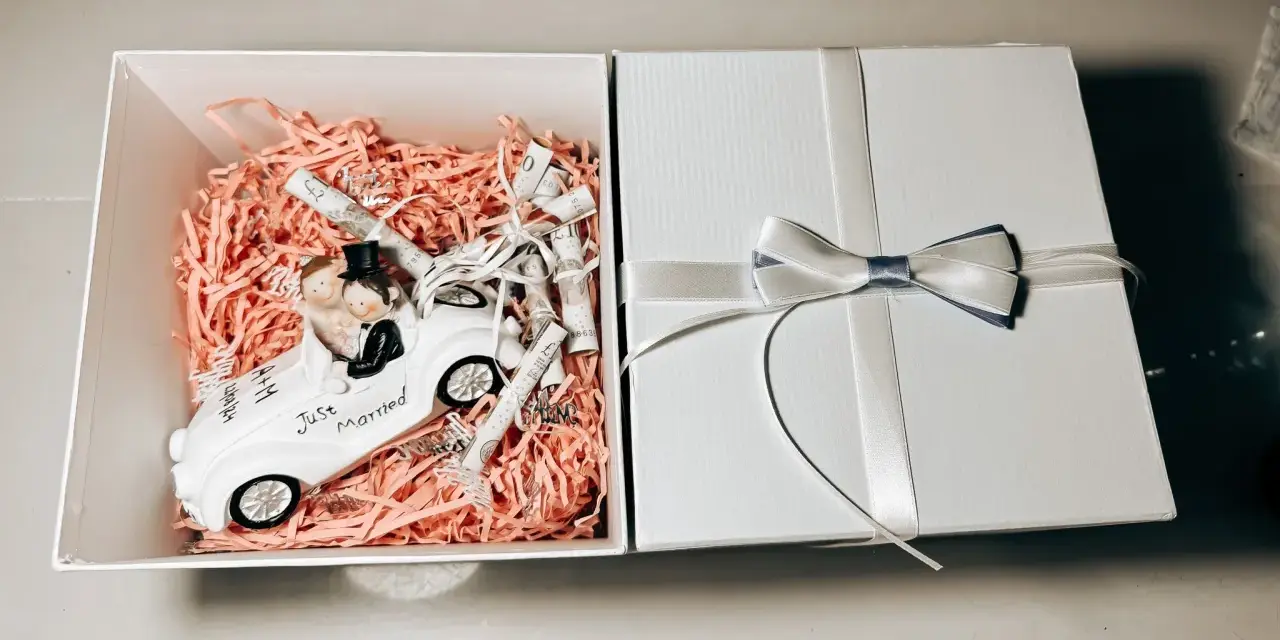 Oto pomysł, jak zapakować zdrapki na ślub: figurka pary młodej w białym samochodzie "Just Married" wśród różowych ozdób i zwiniętych banknotów.