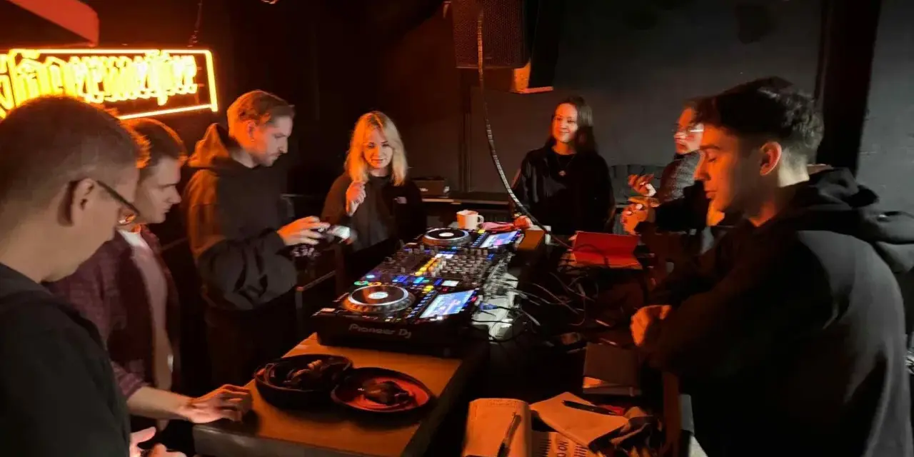 Grupa młodych ludzi przy konsoli DJ-skiej, uczących się, jak zostać DJ-em. Neonowy napis w tle.