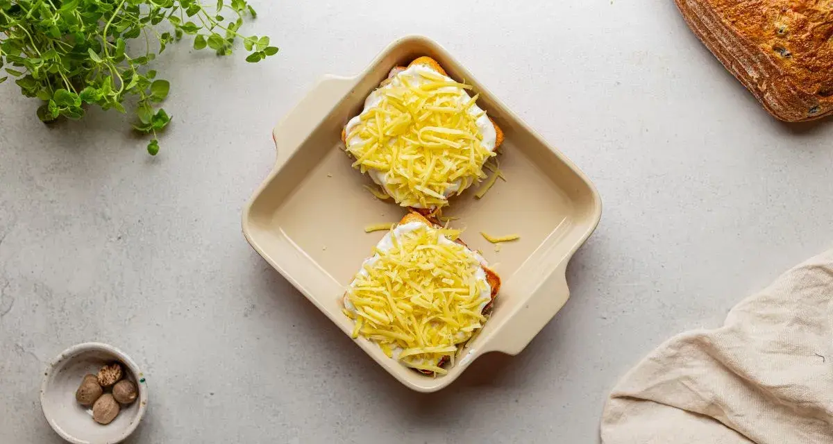 Dwa tosty z serem i beszamelem, przygotowane do zapieczenia jako croque madame. Obok gałka muszkatołowa i świeże zioła.