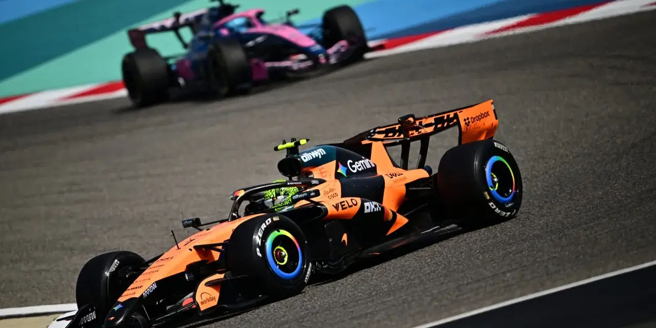 Un coche de Fórmula 1 naranja y negro compite en la pista, anticipando el reglamento F1 2026. Otro coche rosa y azul le sigue de cerca.