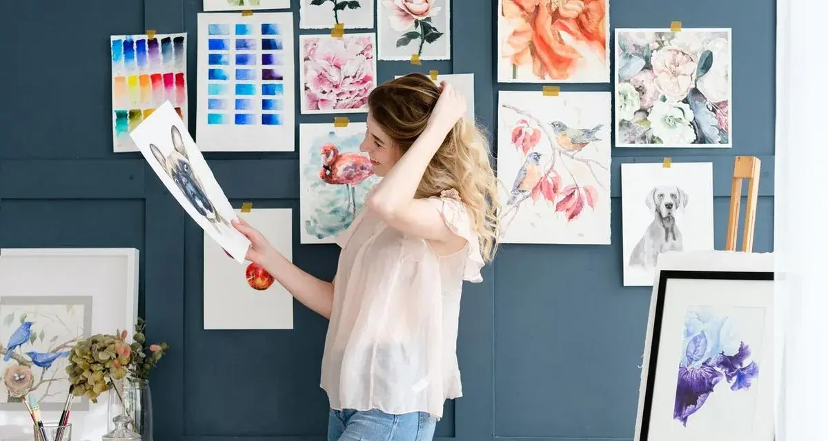 Mujer joven sonríe mientras revisa un dibujo de perro, rodeada de acuarelas de flores, aves y un flamenco. Un espacio inspirador para estudiar dibujo.