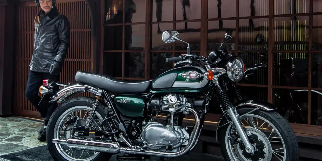 Mężczyzna w skórzanej kurtce i kasku stoi obok klasycznego motocykla Kawasaki W800 w kolorze ciemnozielonym.