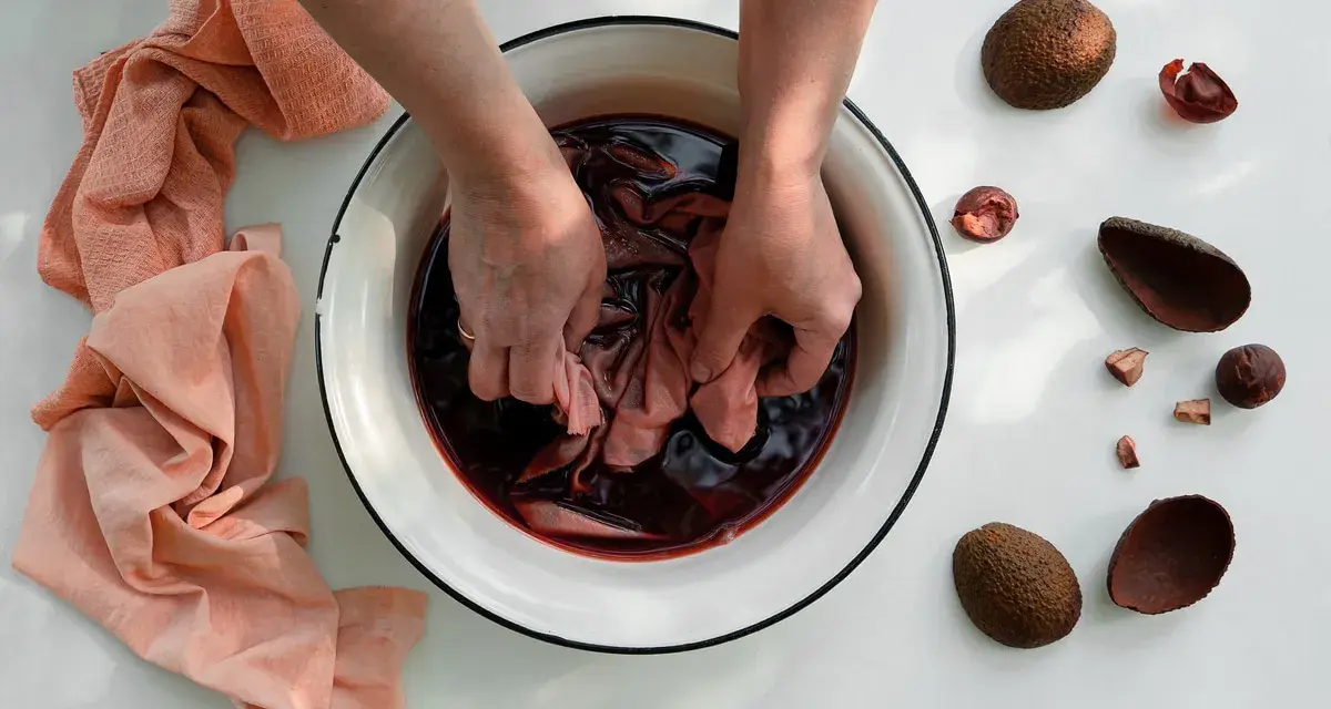 Mani che tingono tessuti in una bacinella con liquido scuro, circondata da bucce di avocado. Un processo naturale per come tingere i tessuti.