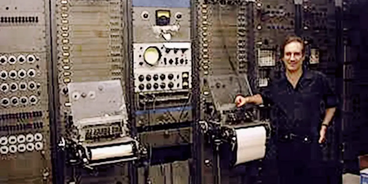 Mann steht neben einem riesigen, frühen Computer, dem ersten Synthesizer der Musikgeschichte.