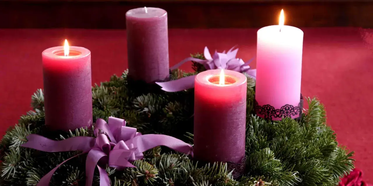 Drei brennende Kerzen auf einem Adventskranz aus Tannengrün mit lila Schleifen. Die dritte Kerze leuchtet hell.