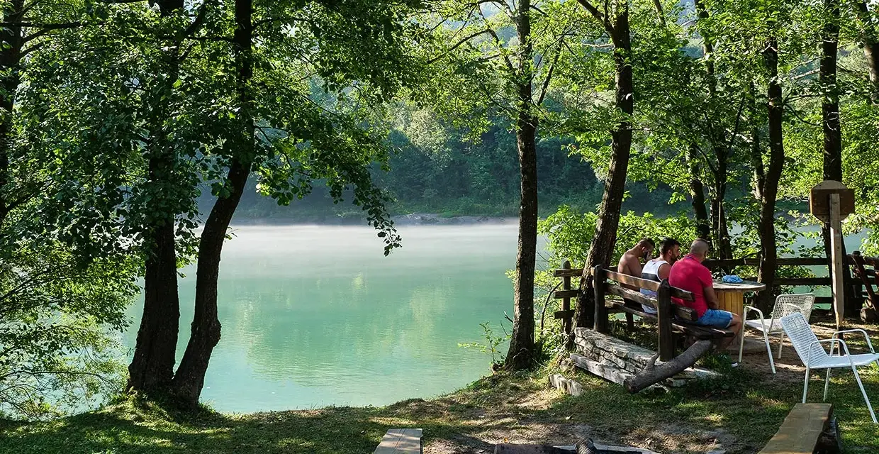 Spokojny poranek na polu kempingowym nad jeziorem. Mgła unosi się nad wodą, a przy stole siedzi kilku mężczyzn.