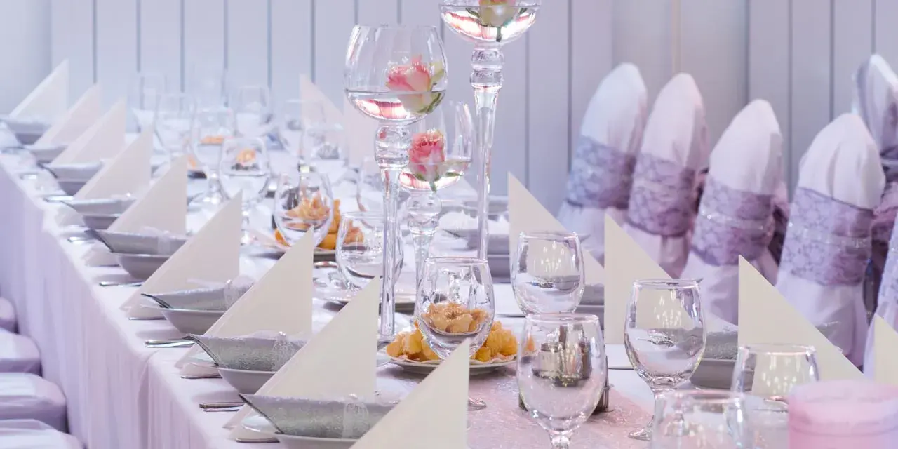 Vacker dukning med rosa rosor i glasvaser, vita servetter och silverfärgade skålar på en rosa bordsduk.