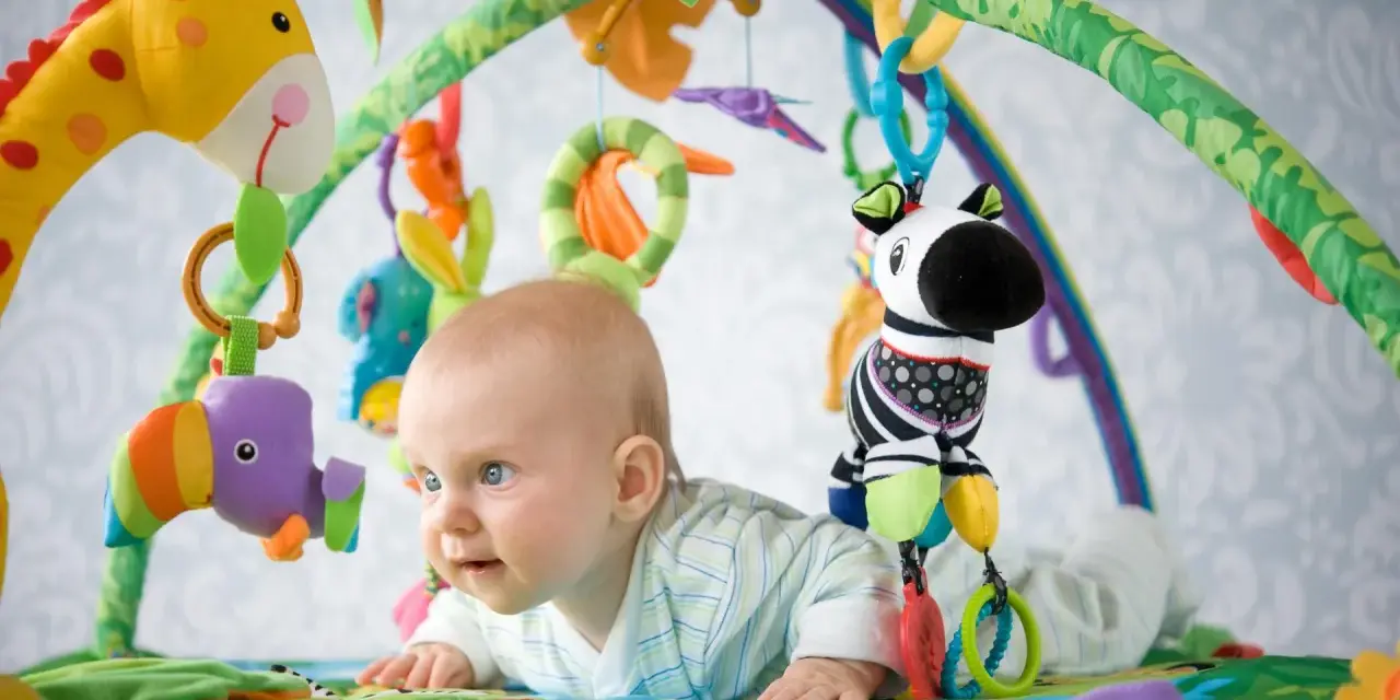 Niemowlę leży na kolorowej macie edukacyjnej, bawiąc się wiszącymi zabawkami: żyrafą, papugą i zebrą.