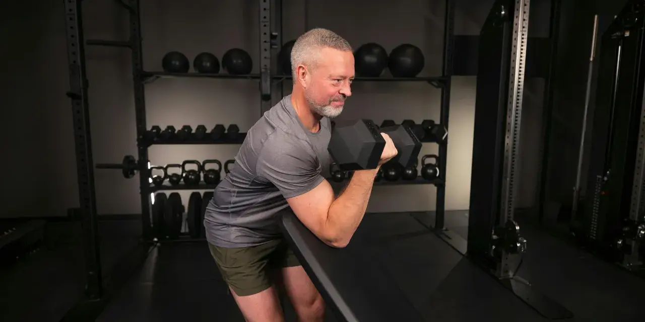 Man med grått hår och skägg utför preacher curls med en hantel i ett gym.