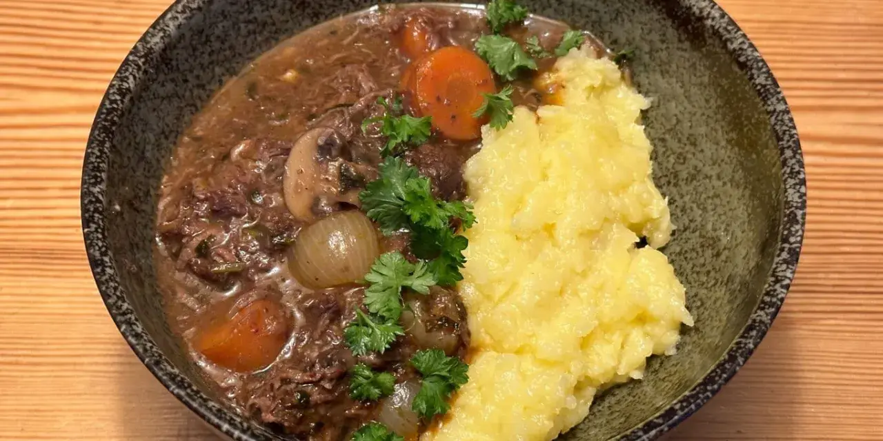 En skål med mustig biff bourguignon med möra köttbitar, champinjoner, lök och morötter, serverad med krämigt potatismos och garnerad med persilja.