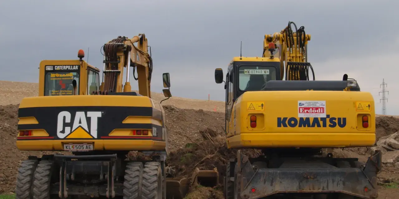 Dwie żółte koparki, CAT i Komatsu, stoją na placu budowy. Czy koparka ma dowód rejestracyjny? Na tablicy rejestracyjnej CAT widnieje KL 505 AE.