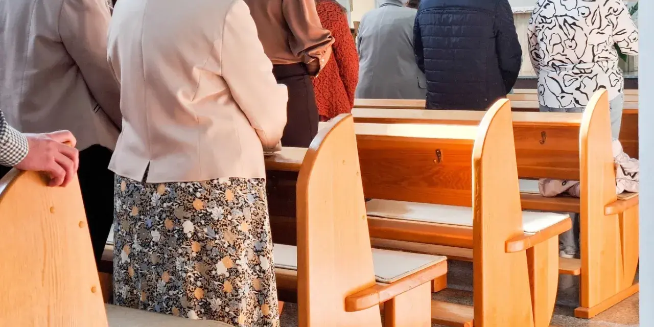 Ludzie stoją w kościele, czekając na dyspensę. Widać ławki i fragmenty ubiorów wiernych.