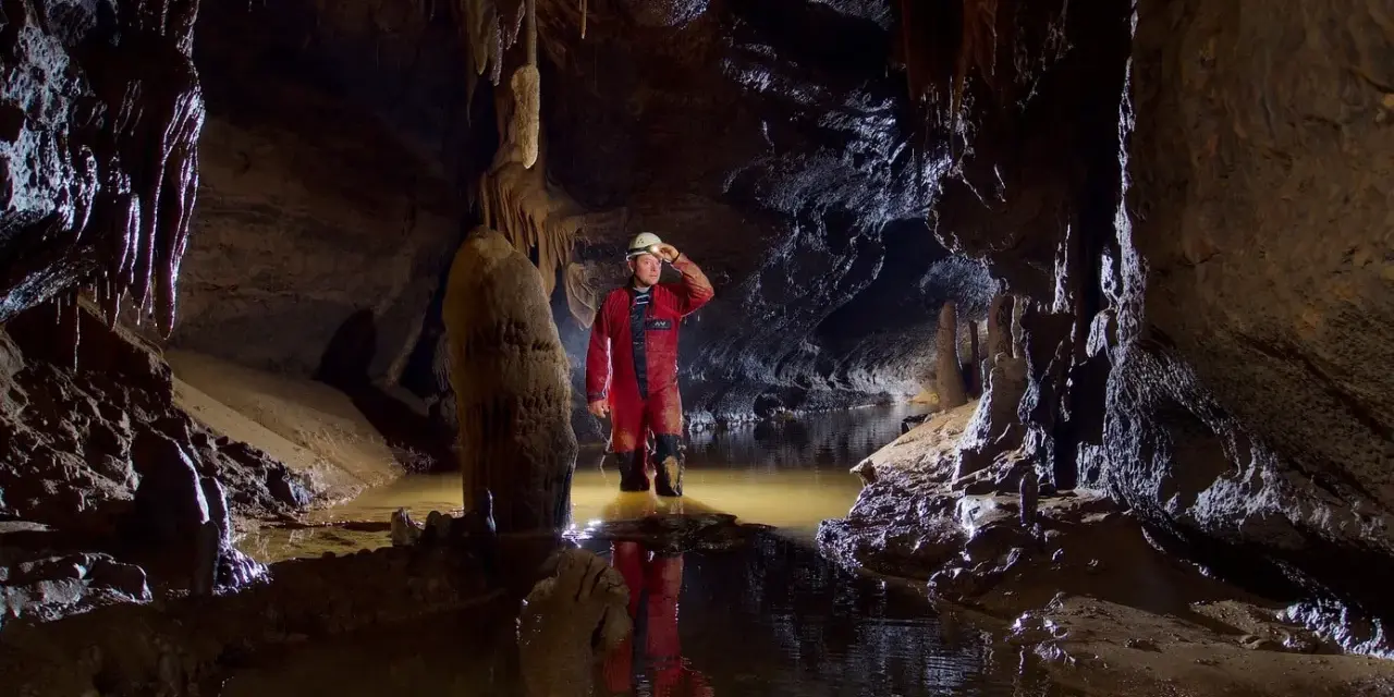 Eksplorator w jaskini, zanurzony po kolana w wodzie, oświetla drogę czołówką. To właśnie jest speleologia – odkrywanie podziemnego świata.