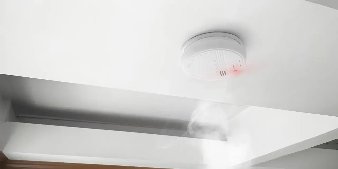Detector de humo blanco en techo, emitiendo luz roja y humo. Cumpliendo la normativa de detectores de humo para seguridad.
