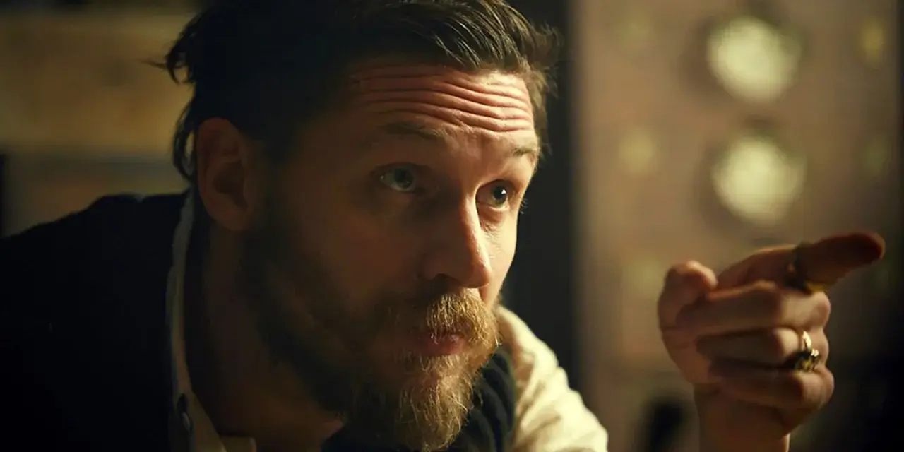 Mężczyzna z charakterystyczną fryzurą w stylu Peaky Blinders, z zarostem i pierścieniami na palcach, wskazuje palcem.