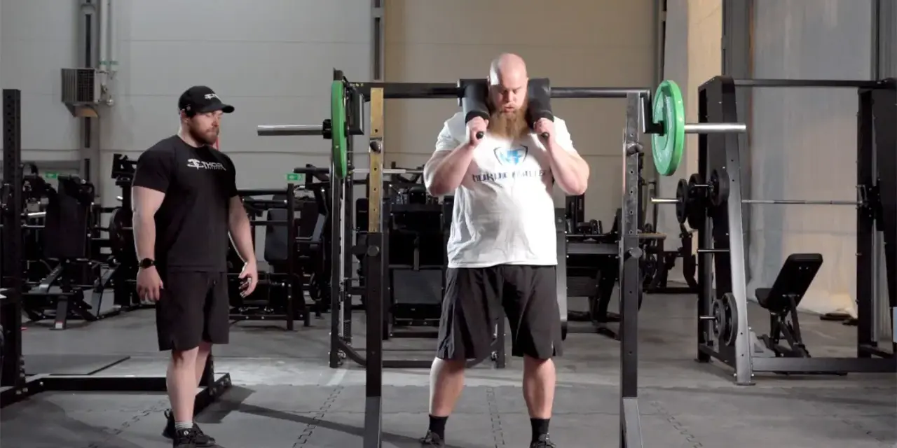 Man med skägg förbereder sig för knäböj med en safety squat bar. En tränare står bredvid.