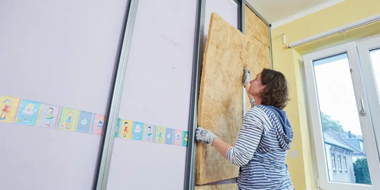 Frau baut eine Trockenbauwand, setzt Dämmmaterial in Metallständer ein.