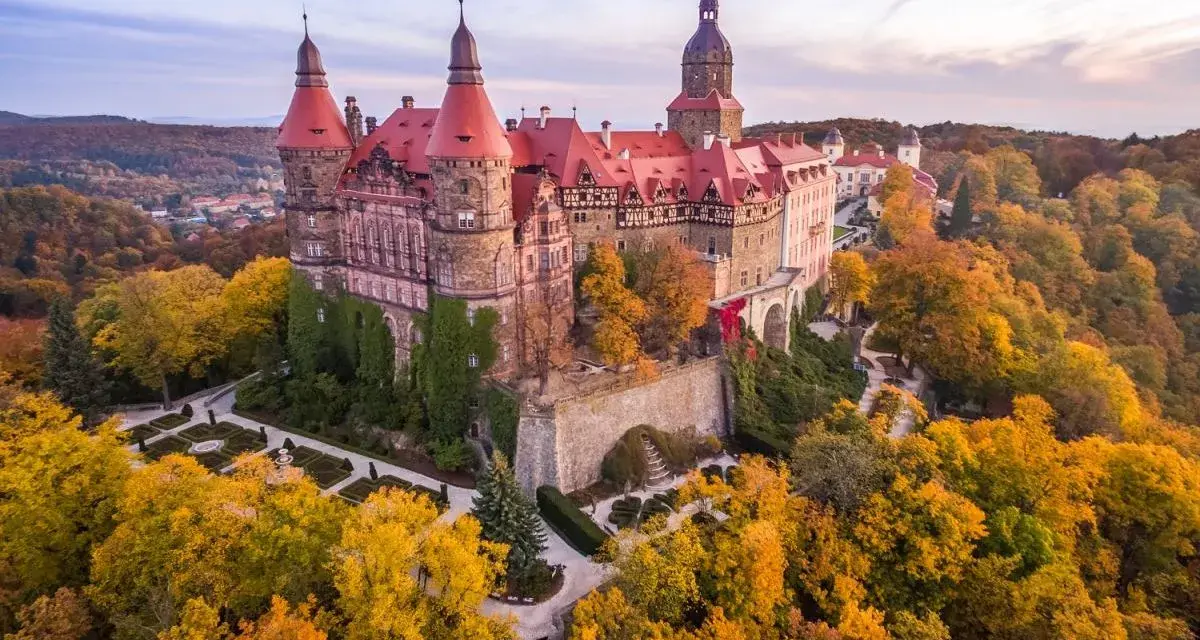 Zamek Książ otoczony jesiennym lasem, z widoczną palmiarnią w oddali.